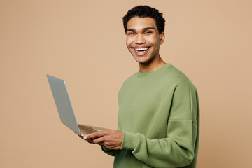 Side view young IT man of African American ethnicity wear green sweatshirt hold use work on laptop pc computer isolated on plain pastel light beige background studio portrait People lifestyle concept