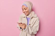 Indoor Shot Of Arabian Woman Wears Hijab And Winter Coat Uses Mobile Phone For Chatting Online Smiles Pleasantly Stands Against Pink Background. Positive Religious Female Model Scrolls Social Networks