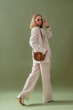 Fashionable Confident Woman Wearing Elegant White Suit With Blazer, Wide Leg Trousers, Trendy Sunglasses, Brown Suede Shoulder Bag, Posing On Green Background. Full-length Studio Fashion Portrait