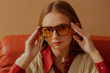 Fashionable confident woman wearing trendy yellow glasses with leopard frames, sitting in armchair, posing on beige background. Close up portrait. Copy, empty space for text