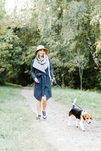 Young Woman In A Coat And Hat Walks A Beagle Dog In The Park
