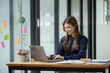 © SOMKID - Happy smiling young asian business woman using laptop sitting at desk in the office, Company worker woman.
