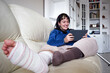 © Mnica - Young woman on couch at home with crutches and orthopedic watching series on tablet. Fracture of the broken leg foot or knee. Concept of rehabilitation and healing. Orthopedics and Traumatology.