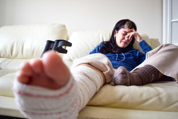 Young woman frustrated tired on couch at home with crutches and orthopedic plaster crying. Fracture of the broken leg foot or knee. Concept of rehabilitation and healing. Orthopedics and Traumatology.