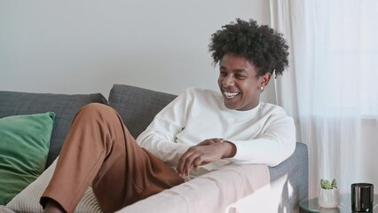 Wall Mural - Young relaxed smiling cheerful happy African American ethnic generation z guy having fun feeling joyful at home laughing looking at camera sitting on couch in modern cozy apartment living room.
