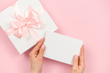 Female hands holding blank white gift card over pink background with gift box, top view