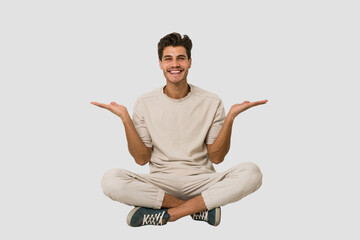 Young caucasian man sitting on the floor isolated on white background makes scale with arms, feels happy and confident.