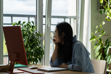 Thoughtful young Asian woman sits at desk with computer trying to come up with cool resume for job search. Procrastinating Japanese girl working from home office gets distracted due to lack motivation