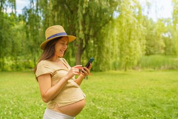 Wall Mural - Pregnant woman using phone app reading about pregnancy or shopping baby things online. Portrait of happy smilling expectant mother in 3rd trimester in city park nature. Asian girl showing tummy belly