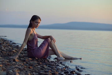 Beautiful young woman sitting on shore of lake in summer dress at sunset. Portrait of a romantic wet girl at sunset, warm sun, natural beauty of a woman