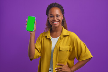 Young smiling casual African American woman demonstrates phone with green screen and keeps hand on belt advertising new trendy mobile application or website stands in lilac studio. Smartphone template