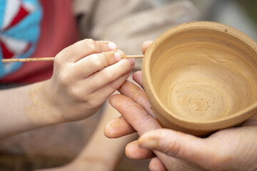 Close-up of a child's hands making a pattern on a clay bowl using a thin stick.A master teaches a child to make pottery on a potter's wheel