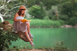 © _KUBE_ - Children's education. Smiling schoolgirl in a dress and a straw hat reading a book sitting on a old log. In the background are park and river. Copy space. The concept of school holidays and curiosity