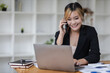 © Wasan - Businesswoman in office working with laptop and documents on desk while talking on phone with customer.