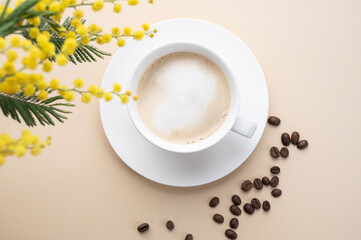 Wall Mural - A spring bouquet with yellow mimosa flowers over a cup of coffee  cappuccino on a beige table with coffee beans. Concept of 8 March, happy women's day.