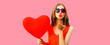 Portrait of beautiful young woman with big red heart shaped balloon blowing her lips sending sweet air kiss wearing sunglasses on pink background
