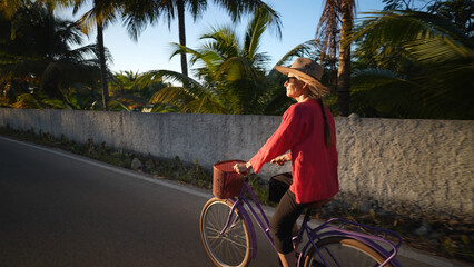 Wall Mural - Moody rim light view of pretty mature senior woman smiling, riding bicycle into the sun past palm trees, wearing sunglasses and a hat. Adventure is ageless.