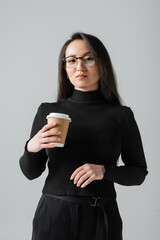 young asian woman in stylish black turtleneck and glasses holding paper cup isolated on grey