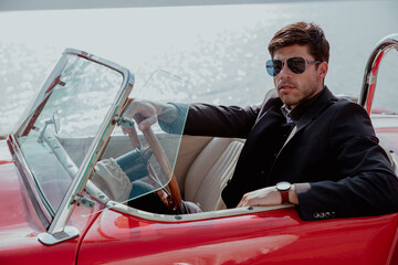 young man in convertible car looking at camera, smiling, man wears suit and tie drives red convertible car