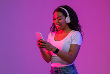 Excited Black Young Woman Wearing Wireless Headphones Using Smartphone In Neon Light