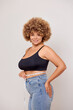 © South House Studio - Self care and weight loss concepts. Vertical shot of half turned cute young woman keeps measurable tape smiles gladly feels confident in her body wears black underlinen bra and casual jeans isolated