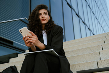 Wall Mural - Young brunette businesswoman using cellphone and laptop outdoors