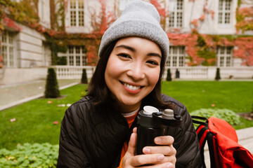 Wall Mural - Cheerful asian woman drinking hot tea from thermos while sitting on bench in old city