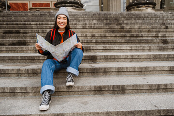 Wall Mural - Positive asian girl tourist examining paper map while sitting on stairs