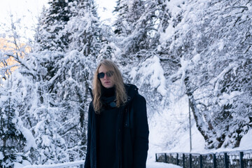 Portrait of model man wearing sunglasses  in winter forest in the mountains, bridge over mountain river, trees covered with snow on slope