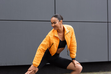 Wall Mural - cheerful african american sportswoman in bike shorts and yellow puffer jacket exercising outside.