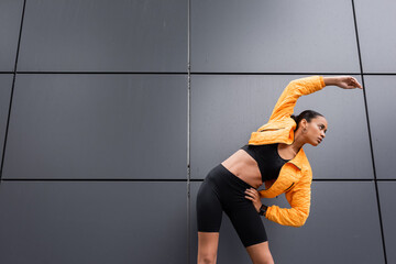 Wall Mural - brunette african american sportswoman in bike shorts and yellow puffer jacket exercising with hand on hip.