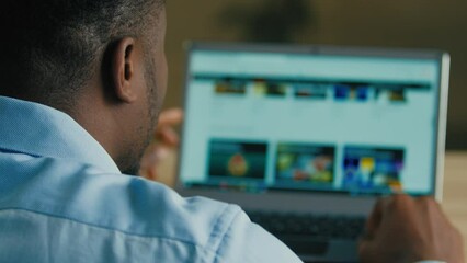 Backside unknown man african american businessman web designer employee sit at home office searching films TV series on internet website e-service looking at computer laptop screen over shoulder view