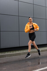 Wall Mural - full length of cheerful african american sportswoman in bike shorts and yellow puffer jacket exercising outside.