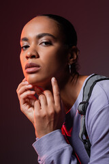 Wall Mural - young african american woman in violet sweatshirt and vest looking at camera on dark purple.