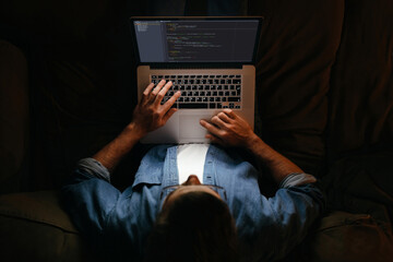 Young coder in eyeglasses is working remotely from home late at night, looking on display, typing code. Hard worker, programmer working overtime, taking work at home, has deadline. 
