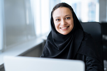 Wall Mural - Beautiful woman with abaya dress working on her computer. Middle aged female employee at work in a business office in Dubai. Concept about middle eastern cultures and lifestyle