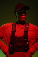 Wall Mural - african american woman in sweatshirt and vest posing with hands on hips on green with red light.