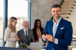 © Dorde - Happy confident handsome wealthy businessman leader holding mobile phone looking at camera standing in modern office for waist up shot photo. Diverse business team defocused in background.
