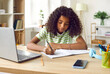 © Studio Romantic - Diligent school child studying at home. Pretty African American girl sitting at desk, doing homework, writing essay in her notebook, using modern laptop and learning a lot of new information