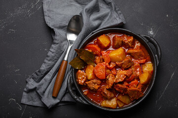 Beef meat stew with potatoes, carrot and delicious gravy in black casserole pot with bay leaves with spoon on black dark rustic concrete background from above .