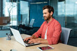 © Liubomir - Distance Learning. Portrait of a young male student in a red shirt. Sitting at the desk in the office, studying online on a laptop. Writes a diploma, a course, an abstract, a lecture.