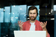 © Liubomir - Distance Learning. Portrait of a young man teacher in a headset in a red shirt sits in the office at the table, works on a laptop online, teaches through a video call. Explains with his hands, smiles