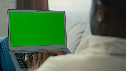 Over shoulder shot african american man businessman programmer freelancer typing on laptop work remote at home on couch looking at green mock-up screen chroma key browsing website make order online