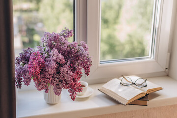 Bouquet of lilacs in a vase,cup of coffee and books on the windowsill.