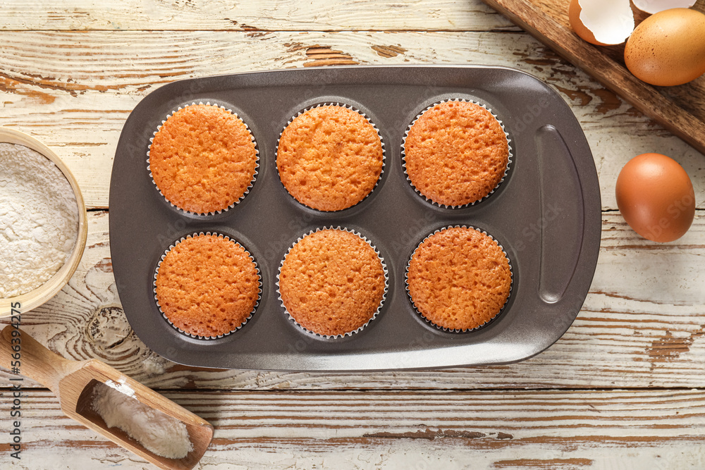 Tray with delicious muffins, flour and eggs on light wooden background