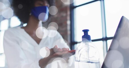 Poster - Animation of light spots over african american woman wearing face mask and disinfecting hands