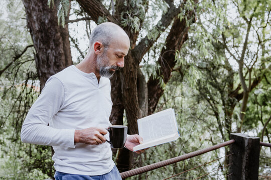 Hispanic man reading a book in the morning and holding coffee cup on the terrace of a country house in Mexico Latin America
