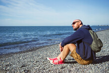 Man In Sunglasses With A Backpack, In Shorts And Red Sneakers Is Resting Alone On A Pebbly Beach By The Sea On A Sunny Day. Background Of Vacation, Relaxation, Freedom