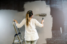 Woman Climbed A Ladder And Catching A Paint Roller Full Of Grey Painting On A Brown Wall. Painter Is Upping And Downing Roller Covering The Wall With Grey Painting What Remains Wet