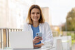 © Maria Vitkovska - Confident smiling young woman holding mobile phone, using laptop computer looking at camera, communication online sitting in modern street cafe. Technology concept